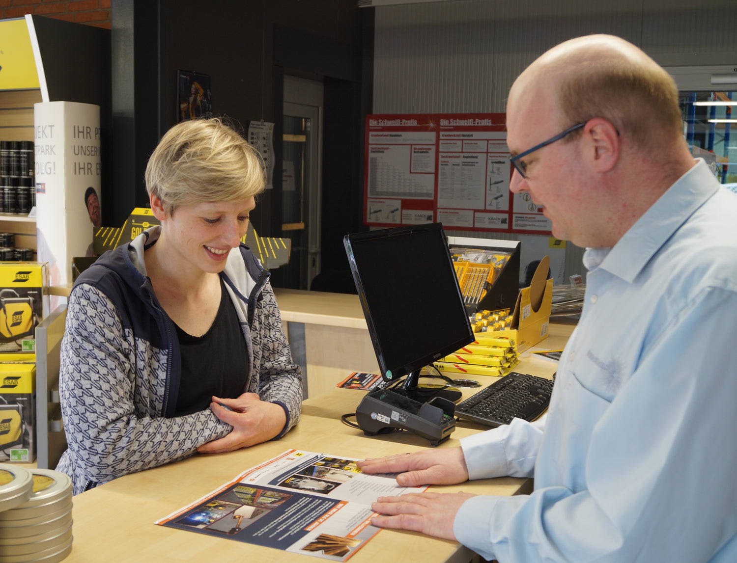 Eine Frau und ein Mann stehen an einem Verkaufstresen in einem Fachgeschäft; der Mann zeigt auf eine Informationsbroschüre, während die Frau aufmerksam zuhört und lächelt. Im Hintergrund sind Verkaufsregale mit Produkten, ein Monitor und mehrere Infotafeln an der Wand zu sehen.