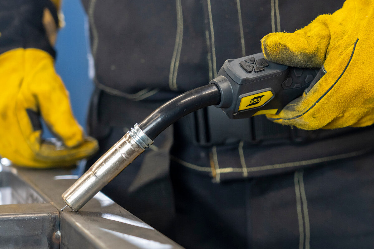 Eine Person trägt schwere Schutzhandschuhe und hält einen MIG/MAG-Schweißgerät-Schlauch in der Nähe eines Metallstücks. Im Hintergrund ist eine metallische Oberfläche sowie eine Arbeitsumgebung sichtbar.