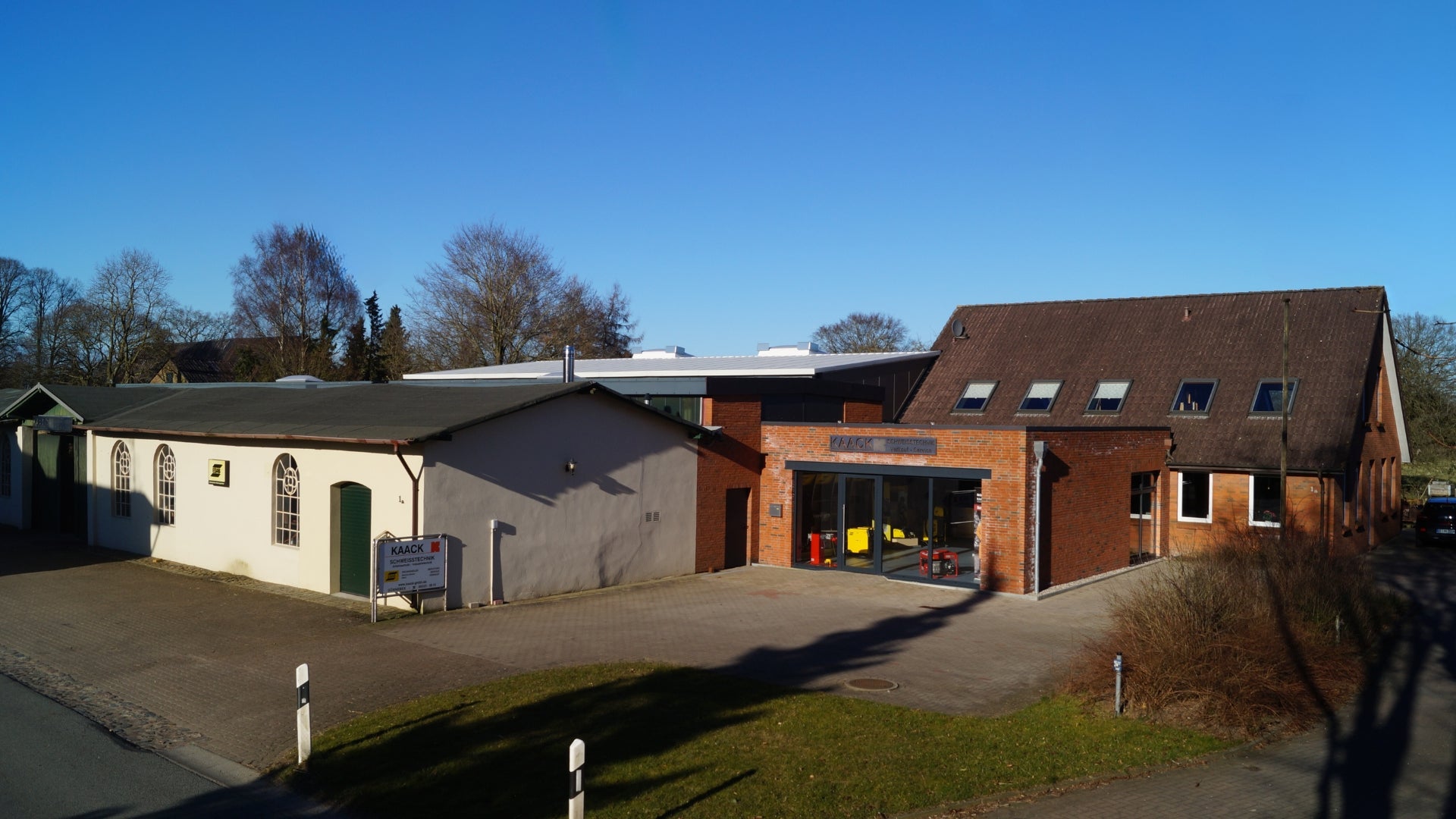 Außenaufnahme eines Firmengebäudes bei klarem Wetter; links ein helles, flachgedecktes Lagerhaus mit grünen Türen und ESAB-Schild, rechts ein moderner Backsteinbau mit Glasfront, Schaufenstern und Firmenschild "KAACK Schweißtechnik", umgeben von Bäumen und gepflastertem Vorplatz.
