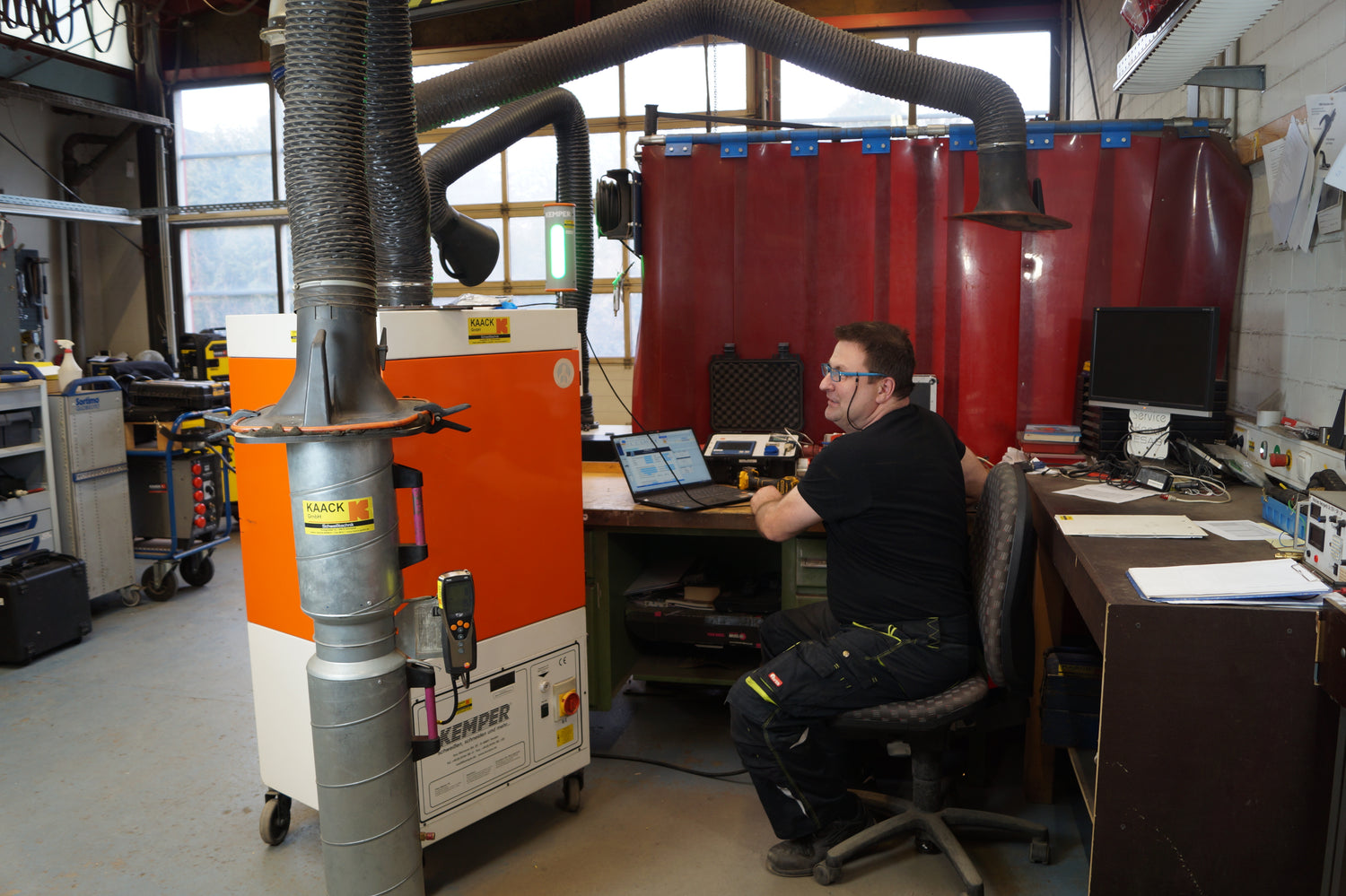 Ein Servicetechniker sitzt in einer Werkstatt an einem Schreibtisch mit geöffneter Diagnosetechnik und Laptop, während im Vordergrund eine mobile Absauganlage mit angeschlossenem Prüfrohr und Messgerät zu sehen ist. Die Umgebung ist mit roten Schutzvorhängen, Werkstattausrüstung und Computerarbeitsplätzen ausgestattet.