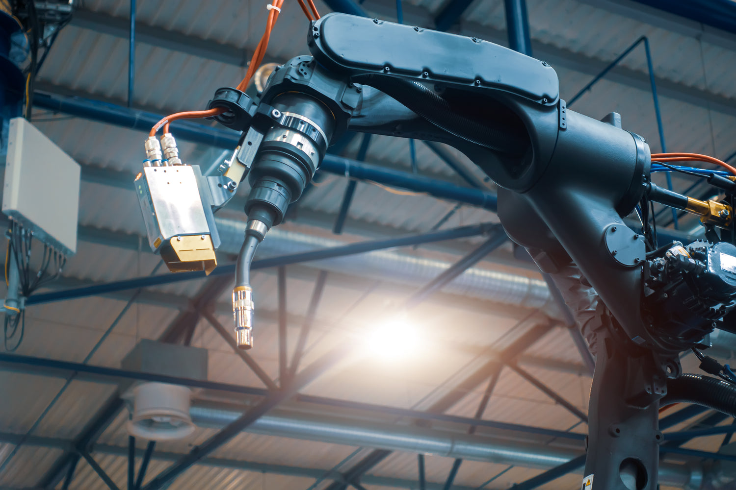 Industrieroboterarm mit angeschlossener Schweißbrenner-Einheit, aufgenommen in einer Werkhalle mit sichtbaren Stahlträgern und Beleuchtung im Hintergrund. Die Schweißtechnik ist mit mehreren Kabeln und Sensoren ausgestattet und bereit für den automatisierten Einsatz.
