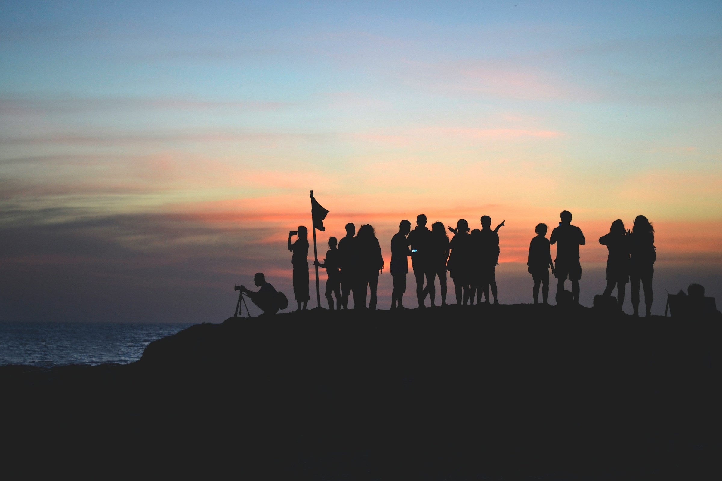 Eine Gruppe von Menschen steht als dunkle Silhouette auf einem Hügel am Meer, während im Hintergrund ein farbenfroher Sonnenuntergang den Himmel in orange, rosa und blauen Tönen färbt. Einige Personen fotografieren, andere unterhalten sich oder schauen in die Ferne.