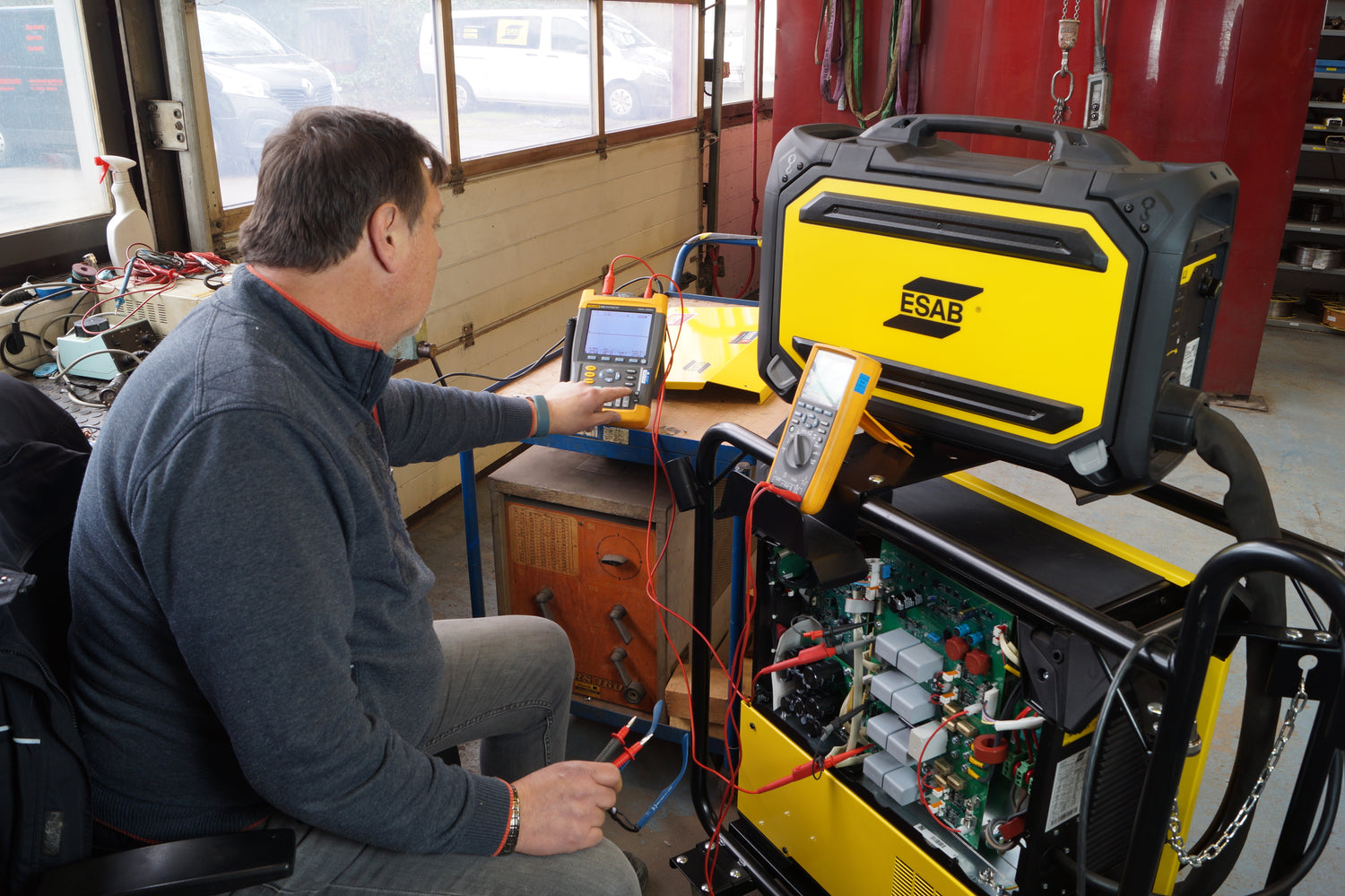 Ein Techniker sitzt in einer Werkstatt an einem geöffneten Schweißgerät mit gelbem Gehäuse und führt mithilfe mehrerer Messgeräte und Prüfleitungen eine elektrische Diagnose durch. Auf dem Arbeitstisch befinden sich zusätzliche Geräte und Kabel, im Hintergrund ist ein Fenster mit Blick nach draußen zu sehen.